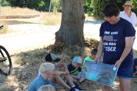 Rechts im Bild ist eine Person die ein aufgeschlagenes Buch mehreren Kindern zeig, die auf dem Boden sitzen. Die Personen sind draußen. Im Hintergrund sieht man den Stamm eines Baums. Die Kinder sitzen in dessen Schatten. Die Wiese ist sehr vertrocknet.